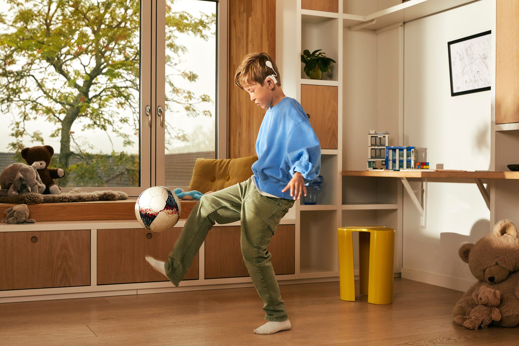 Ung gutt med cochleaimplantat trikser med fotball foran et vindu i en stor stue